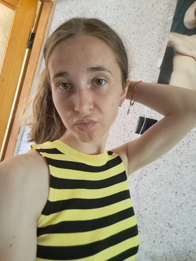 A young woman with light brown hair pulled back in a ponytail, fair skin with freckles, and brown eyes takes a selfie. She is wearing a yellow and black striped tank top. Her right arm is raised behind her head. She is standing indoors with a wooden door frame to her left and a textured wall behind her. A black electrical outlet is visible on the wall to her right. There is a poster partially visible on the wall behind her, showing a person's arm and shoulder. The lighting appears to be natural, coming from the right side of the frame.