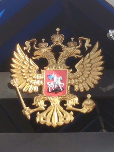 The image shows a close-up, low-angle shot of a golden double-headed eagle crest. The crest is ornate, featuring two prominent crowns atop the eagle's heads, and smaller crowns on the eagle's wings. A red shield with a silver equestrian figure is centrally located on the eagle's chest. The eagle's wings are spread wide, with detailed feathering. A golden scepter with a crown finial is held in the eagle's left talon, and a golden orb topped with a cross is held in the right talon. The crest is mounted against a dark blue, textured background, with diagonal lines suggesting a structure or facade. The lighting is bright, casting highlights and shadows on the metallic surface of the crest, emphasizing its three-dimensional form. The overall impression is one of grandeur and official insignia. This crest is located at the Royal Thai Naval Academy in Ko Kaeo, Thailand.