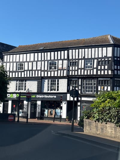 The image shows a three-story Tudor-style building on a street in Nantwich, United Kingdom. The building features a black and white timber-framed facade with white stucco infill. The ground floor of the building houses several businesses, including "D&S Electrical Distributors" and "Mia Store". Storefronts are visible with signage and displays. Several street signs, including a "no entry" sign, are present. A tree is partially visible on the left side of the image. A low stone wall with dense green foliage runs along the right side of the sidewalk. The street in the foreground appears to be paved and has lane markings. The sky is clear and blue. The overall scene depicts a commercial street in a historic town.