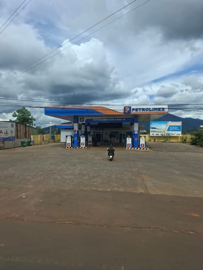 The image depicts a Petrolimex gas station in Tân Phú 2, Vietnam, under a cloudy sky. The gas station, branded with the Petrolimex logo, features a blue and orange canopy over the pumps. There are multiple gas pumps visible, each with screens displaying prices and information. A lone individual on a motorcycle is seen driving away from the pumps towards the foreground of the image. In the background, there are buildings, trees, and mountains. To the left of the gas station, a billboard displays information about insurance. To the right, another billboard advertises Petrolimex products and services. The ground in front of the gas station is paved, with some areas appearing weathered and cracked. The overall scene suggests a typical day at a roadside service station in a semi-rural or suburban area.
