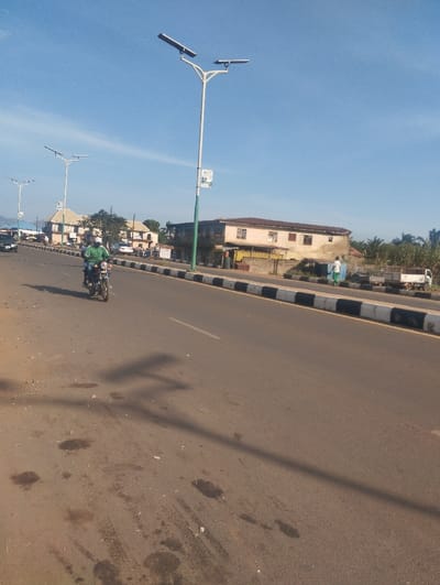 The image displays a roadside scene in Unguwan Rimi, Nigeria, under a clear, light blue sky with no visible clouds. The perspective is low-angle, looking down a multi-lane asphalt road that extends into the distance. The road surface in the foreground exhibits dark stains and scattered debris; long shadows are cast across the asphalt, indicating a low sun angle.
A black and white striped curb separates the main roadway from an adjacent shoulder or unimproved area on the right.
Prominently positioned on the right side of the road is a tall white street light pole with a green base. It features two upward-angled solar panels, each supporting an LED light fixture. A rectangular green and white sign is attached to this pole. Several additional identical streetlights are visible further down the road on the left.
One individual is operating a motorcycle on the left lane of the road, traveling away from the camera. This person is dressed in a light green shirt and dark trousers. A dark-colored passenger car is also visible in the distance on the left lane.
To the right of the road, beyond the curb, stands a two-story building with a light brown/pinkish facade, multiple windows, and a darker roof. Smaller structures are visible at its ground level. Further right, a white utility truck with an open cargo bed is parked. Two individuals are present in this area; one, wearing a light-colored shirt and dark trousers, walks away from the camera near the truck, while a second, less distinct figure is nearby.
In the background, on the left side of the road, several multi-story buildings of varying architectural styles are visible, interspersed with trees and other vegetation. Palm trees are also observed behind the structures on the right side of the scene.