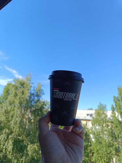 A hand holds a dark disposable coffee cup against a bright blue sky with scattered white clouds. The cup has a black lid and is decorated with small white silhouettes of vehicles, including cars and motorcycles. The logo on the cup reads "il MOTORRE COFFEE" in white text, with a small Italian flag design next to the word "MOTORRE."
In the background, lush green trees with blurred foliage create a soft bokeh effect. Beyond the trees, the top of a building with a light-colored facade is visible. The clear blue sky and bright sunlight suggest it is daytime, likely morning or afternoon. The overall scene evokes a feeling of a pleasant, casual moment outdoors.
The context indicates this photo was taken in Arkhangelsk, Russia.