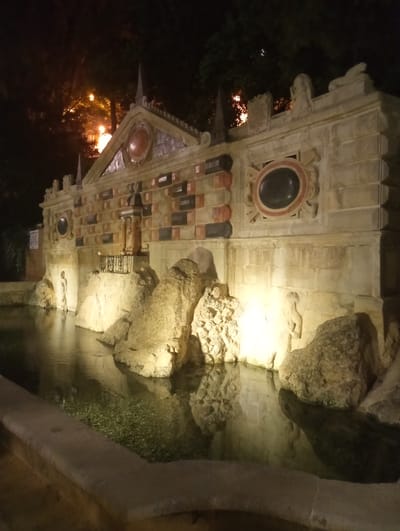 The image shows the Fuente del Rey in Priego de Córdoba, Spain, at night. The fountain is made of stone and features several sculptures and decorative elements. In the foreground, there is a pool of water with rocks and aquatic plants. The fountain itself is a large, ornate structure with a central part that has a tiered design. There are statues of figures, some of which appear to be mythological. The stonework is detailed, with columns, arches, and carved reliefs. The lighting is dramatic, with spotlights illuminating the fountain and creating strong shadows, emphasizing the textures and forms of the architecture. Trees are visible in the background, silhouetted against the dark night sky. The overall impression is one of grandeur and historical significance.