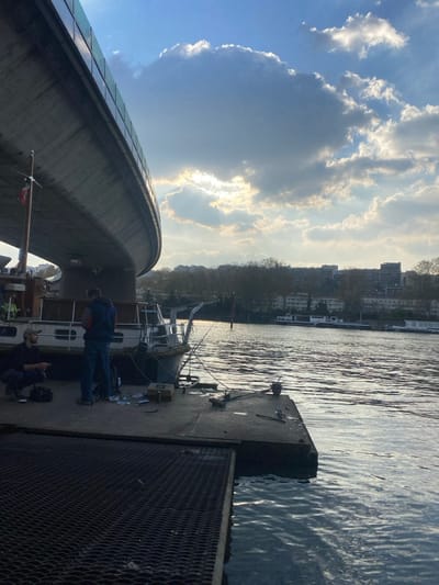 The image depicts a river scene in Boulogne-Billancourt, France. A large, curved concrete overpass or bridge, with a glass-paneled barrier along its upper edge, dominates the upper left quadrant of the frame. Beneath this structure, on a concrete pier, two adult males are present.
The man on the left is crouched, wearing a dark cap, a dark jacket, and dark pants, with a beard visible. He appears to be attending to something on the pier surface, possibly a small bag located beside him. The second man stands centrally, dressed in a dark blue jacket, blue jeans, and dark shoes, facing towards the river or a docked vessel.
A grey-hulled motorboat with a white superstructure is moored alongside the concrete pier, positioned directly under the bridge. Several fishing rods are visible, extending from the boat or propped near it. Various items, including a light brown metallic tackle box, are scattered on the pier next to the boat, indicating active or recent fishing. Further under the bridge, a larger vessel, possibly a houseboat or cabin cruiser, is partially visible, featuring a small mast with a red-and-white flag.
The foreground includes a dark, metal-gridded platform transitioning into the concrete pier. The river water exhibits ripples and reflects the sky, appearing luminous. The sky is partially covered by cumulus clouds, with the low sun obscured but producing a bright glare from behind the clouds in the upper right quadrant. The distant opposite bank of the river displays a line of bare trees, behind which a collection of multi-story urban buildings is visible. Other boats are moored further downstream on the river. The overall lighting suggests late afternoon or early evening.
