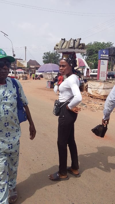 The scene depicts an outdoor urban environment, specifically a street or market area in Igbo Ukwu, Nigeria, under an overcast or hazy sky. The ground is an unpaved or worn asphalt surface.

In the foreground, slightly to the left, an older male is partially visible, facing towards the right. He wears a green baseball cap, dark sunglasses, and a light-colored short-sleeved shirt patterned with blue geometric squares. A blue backpack is slung over his left shoulder. His left arm and hand are visible, and he wears sandals.

Centered in the mid-foreground is a female subject, standing in profile, facing right. She has dark skin, wears a white long-sleeved top, black fitted pants, and dark grey slide sandals. Her toenails are painted white. Her head is covered by a patterned headscarf, predominantly black with white starburst or floral motifs. She carries a small black cross-body bag with a white "LV" style logo visible on its front. Her hands are clasped behind her back.

To the immediate right of the female, a portion of another individual's arm and hand is visible, holding a dark, possibly plastic, bag or object.

In the mid-ground and background, a bustling street scene unfolds. To the left, a street light pole is present. Further back, a light-colored van or small bus is parked. A brown rectangular sign with illegible text is mounted on a pole or building. A purple umbrella, indicative of a vendor stall, is visible.

To the right, a prominent blue sign with white lettering reads "CAPAD COMPANY NIG. LTD." and below that, "FUEL KERO DIESEL," indicating a fuel station. Above the female subject and the "CAPAD" sign, a decorative horizontal platform features several uniformly shaped, light-colored rectangular objects resembling stylized jerrycans or oil drums. Green foliage from trees is visible in the background. Overhead, power lines traverse the sky. Scattered debris and what appear to be bundled goods are visible on the ground near the fuel station sign. Several other individuals are visible in the background, walking or attending to market-like activities.