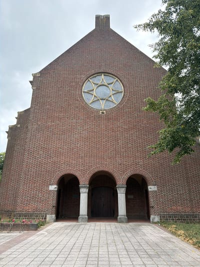 The image shows the facade of a brick church in Melick, Netherlands. The church has a large rose window with a star pattern above a set of three arched entrances. The entrances are supported by stone columns. A tree branch with green leaves extends into the frame from the right side of the image. The ground in front of the church is paved with grey square tiles. In the lower left corner, there are some pink flowers and a bicycle rack. The sky is overcast.