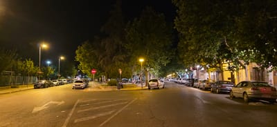 The image depicts a street scene at night in Huesca, Spain, likely on the outskirts or a residential area with a park. The street is lined with trees casting shadows and is illuminated by streetlights. Several cars are parked along both sides of the road. On the left side of the road, a fence and some green foliage are visible. There's a circular area with a no-entry sign and a small park or garden in the center of the intersection. On the right side of the street, buildings with storefronts are visible. The road has traffic markings, including directional arrows. The sky is dark, indicating nighttime.