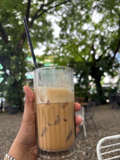 In Bireuen, Indonesia, a person's hand, adorned with a rose gold and silver watch, holds a tall, fluted glass filled with iced coffee. The drink, a light brown color with a frothy top, contains ice cubes. A black straw extends from the top of the glass. The background is blurred, revealing the dappled sunlight filtering through green leaves of trees and the out-of-focus shapes of outdoor seating and a gravel ground. The scene suggests a relaxed outdoor cafe setting.