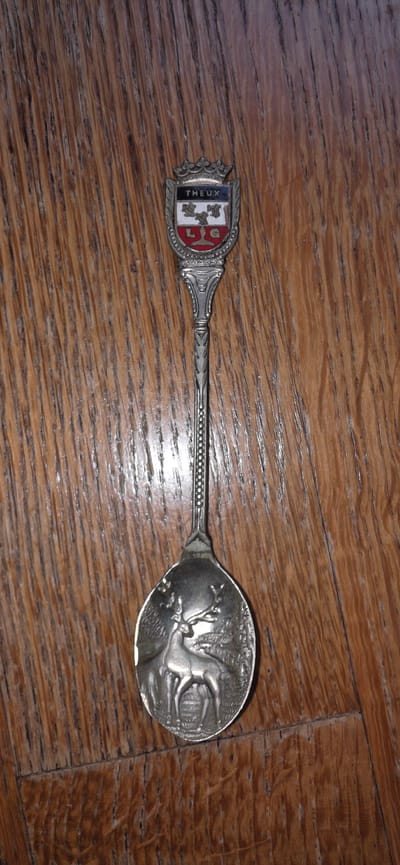 A detailed, full-shot, high-angle, vertical image shows a single ornate silver spoon lying on a wooden surface. The spoon has a decorative crest at the top with the word "THEUX" written in black lettering. Below the crest is a coat of arms with red and white sections and a depiction of a tree or plant. The handle of the spoon is intricately patterned, and the bowl of the spoon features a raised relief of a deer in a forest setting. The wooden surface appears to be made of planks with a visible grain. The location in the city of Marum, Netherlands is not identifiable from the image.