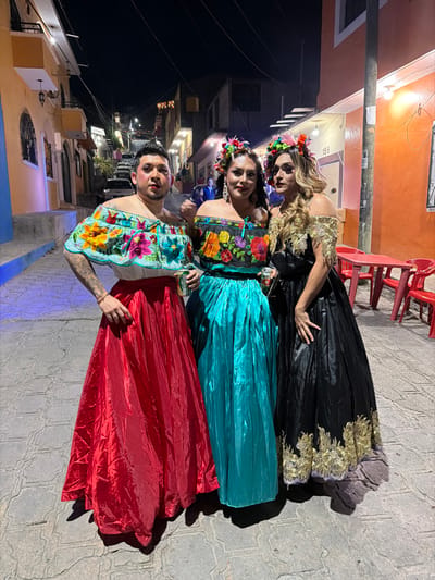 Three individuals in vibrant traditional Mexican dresses pose on a dimly lit street. The dresses, colored black, teal, and red, are adorned with intricate floral embroidery and shimmering fabrics. All three wear floral headbands. The background reveals a mix of buildings and street lights, adding to the festive atmosphere.