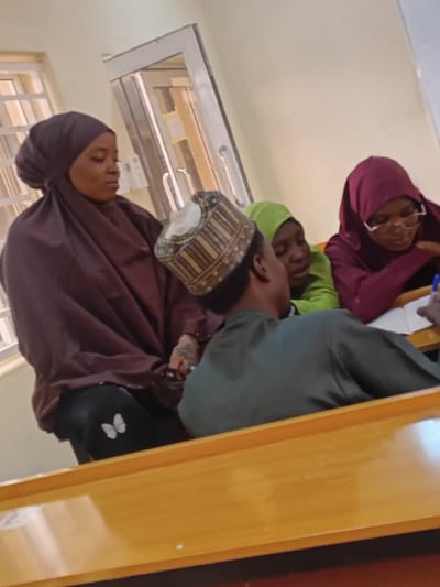 An indoor scene in Kwazailewa, Nigeria, shows four individuals seated at light-colored wooden desks. In the foreground, a male individual, viewed partially from behind, wears a dark green long-sleeved garment and a patterned cap; he holds a pen poised over a notebook. To his immediate left, a female wearing a dark brown hijab and matching attire is seated, observing the group; visible tattoos are present on her left forearm, and she wears black leggings with a white butterfly motif on the left leg. Behind the foreground male, a second female in a light green hijab is partially visible. To the right of the foreground male, a third female, wearing a dark red hijab and glasses, is focused downwards on written material. The background consists of light-colored walls, a barred window on the left, and a glass-paned door on the right.