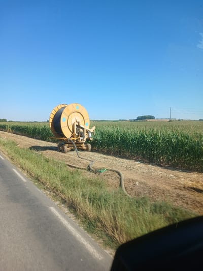 In Cléry-sur-Somme, France, a bright yellow irrigation reel sits stationary on a dirt path next to a field of tall green corn. A thick, dark hose snakes from the reel and curves across the ground, disappearing into the vegetation. The reel itself is mounted on a wheeled trailer, suggesting it's a piece of farm equipment. The sky above is a clear, uninterrupted blue. The scene appears to be a rural setting, likely agricultural land.