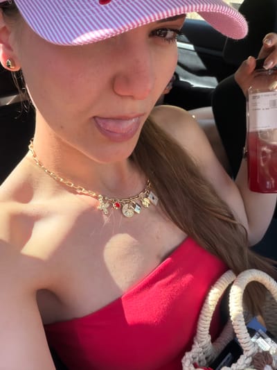 A young woman wearing a pink and white striped visor and a strapless red top is shown in a car. She has a gold chain necklace with various charms, including a bee, a red gem, a compass, and a lock. She is sticking her tongue out and holding a bottle of red liquid. A portion of a woven bag with rope handles is visible. The lighting suggests it is daytime. The scene is not specifically indicative of Kyustendil, Bulgaria.