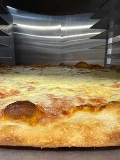 The image shows a close-up view of a pizza being cut by a pizza cutter. The cutter has multiple sharp, triangular blades made of reflective metal. The pizza has a thick crust and is covered with cheese and what appears to be sauce. The crust has a golden-brown color, and some areas of the cheese are slightly browned. The background is out of focus. The location, West Ocean City, United States of America, is not discernible from the image.