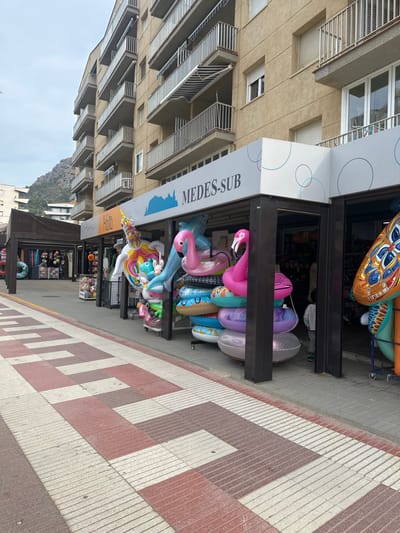 The image captures a commercial street scene in L'estartit, Spain. A wide pedestrian promenade dominates the foreground, paved with alternating light-colored and reddish-brown rectangular slabs, some of which feature ridged textures. To the right, a series of open-fronted commercial kiosks are situated beneath a multi-story building with a light beige facade and numerous balconies equipped with metal railings; some balconies display striped awnings.

The most prominent kiosk, identified by a white sign bearing the blue text "MEDES-SUB" and a silhouette of islands, displays a dense collection of inflatable pool floats. These include two large pink flamingos, a white unicorn, a light blue dolphin-shaped float, and various colorful circular swim rings, all arranged on stands within and outside the storefront. To the immediate left of "MEDES-SUB," another kiosk with a partially visible sign, possibly reading "Maria" or "Marta," also exhibits merchandise.

A single small child, viewed from the back, is positioned near the right entrance of the "MEDES-SUB" store, wearing light-colored clothing and green shoes. No other individuals are clearly visible. In the background, beyond the buildings on the left, a mountainous landscape is visible under an overcast sky. The overall lighting suggests a cloudy day.