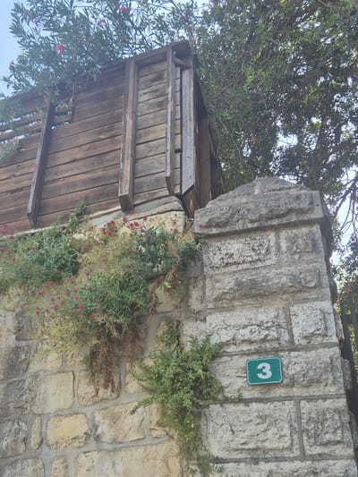 The image depicts a section of a stone wall in Abu Gosh, Israel, adorned with a green house number sign displaying the numeral "3." Above the wall, a wooden structure with horizontal planks is visible, partially obscured by lush green foliage and small pink flowers that cascade down the stone facade. Trees with dense green leaves overhang the scene. The stonework of the wall is rough-hewn and appears aged.