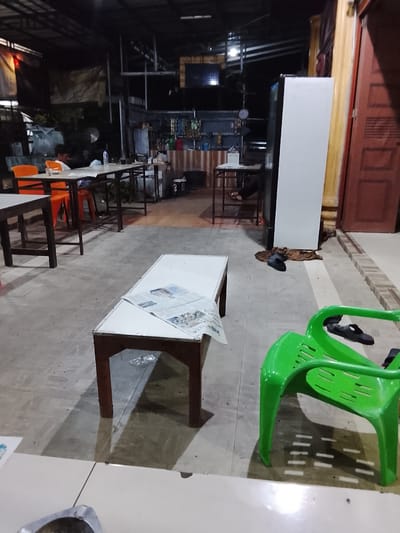 The image depicts an outdoor seating area at night, likely a food stall or small restaurant in Dayahlhoksukon Barat, Indonesia. The scene is dimly lit, with artificial lights illuminating the tables and chairs. In the background, there's a structure with a corrugated metal roof, suggesting a covered outdoor space. Various items are visible, including tables, chairs, a large white refrigerator, and what appears to be a newspaper on one of the tables.
There are at least two people in the image, though their faces are not clearly visible. One person is seated at a table in the background, appearing to be engaged in eating or talking. Another person's legs and feet are visible under a table in the mid-ground, indicating they are also seated. A bright green plastic chair is prominent in the foreground, with a pair of black sandals on the floor next to it. Another pair of sandals is seen near the refrigerator.
The overall atmosphere is casual and utilitarian. The time of day is clearly night, indicated by the artificial lighting and the dark surroundings. There are no clear indications of weather conditions. The presence of multiple tables and chairs suggests a public space designed for dining or socializing. The newspaper on the table suggests a place where people might relax or catch up on news while dining. No discernible text is visible in the image.