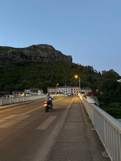 The image shows a bridge over a river in Anduze, France at dusk. A motorcycle is visible on the bridge, with a rider on it. The bridge has a pedestrian walkway and a road with painted lane markings and a crosswalk. Buildings are visible on the left side of the bridge, and a hill covered in trees is in the background. A street lamp is lit on the right side of the bridge. The sky is clear and a pale blue.