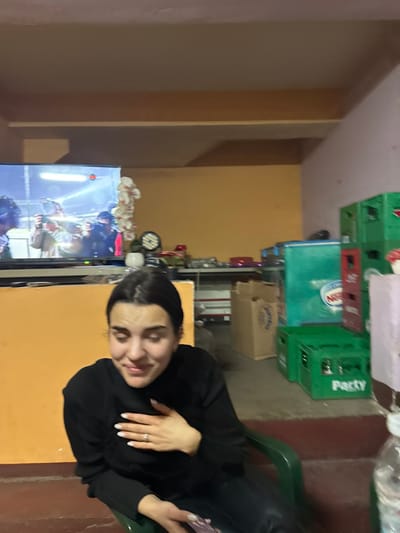 The scene depicts an indoor setting, likely a casual living or storage area, in Gorna Grashtitsa, Bulgaria. A female individual with dark hair, wearing a black long-sleeved top and dark pants, is seated in a green plastic chair in the foreground, angled towards the right. Her eyes are partially closed, and she displays a smiling or laughing expression, with her left hand placed on her chest, revealing a ring on her ring finger. She holds a white smartphone in her right hand.

To her left, a large flat-screen television displays an image of multiple individuals holding cameras or phones, possibly at a concert or event. A white, floral-like decorative string hangs vertically to the right of the television. Behind the woman and extending to the right, the room contains various objects: multiple green plastic crates are stacked, some bearing the word "Party," alongside red plastic crates and cardboard boxes. A light blue or greenish refrigerator or cooler, partially obscured, shows the word "Nestle." A circular light fixture is visible on a surface behind the television. The walls are painted a distinct yellow-orange on the left and a light pinkish-purple on the right. The ceiling is a beige color with visible beams. The floor is dark, with reddish-brown areas. A plastic water bottle and a pink object are visible in the immediate right foreground.