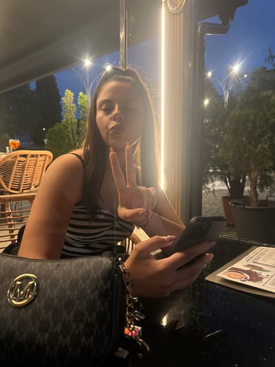 A young female, appearing to be in her late teens or early twenties, is seated at an outdoor cafe or restaurant in Simitli, Bulgaria. She is making a "kiss" face with her eyes closed and displaying a peace sign with her raised left hand. Her right hand holds a dark-colored smartphone. She wears a striped strapless top, and her long, dark hair is styled in a high ponytail. A dark shoulder bag with a prominent gold "M" logo and repeating "M" pattern is visible in the lower left foreground.
To her left, behind her, is a wicker-style chair and a glass containing an orange beverage with a garnish. A dark, possibly scratched, table surface is in front of her, on which a menu featuring coffee beans and a coffee cup is visible to the right. The scene is illuminated by artificial light sources, indicating it is nighttime. A bright, vertical light fixture or patio heater is to her right. The background shows a dark blue sky, streetlights with distinct arched poles, and various trees and potted plants, suggesting an urban outdoor setting.