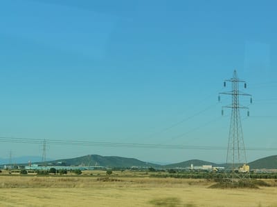 The image depicts a vast, dry, yellow field stretching towards a backdrop of rolling green hills. A clear blue sky dominates the upper portion of the frame. In the mid-ground, a large industrial complex with low-lying buildings and several tall structures is visible. Two prominent electricity pylons stand in the foreground and mid-ground, with power lines crisscrossing the scene. The overall impression is one of a rural industrial landscape under a bright, sunny sky. The location is the Municipal Unit of Almyros, Greece, a region known for its agriculture and industrial development.