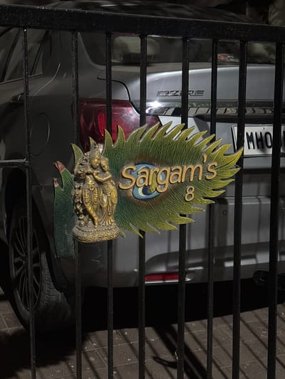The image presents a static scene through a black metal gate in a residential or private outdoor area in Ambernath, India. A decorative nameplate is affixed to one of the vertical bars of the gate. This nameplate consists of a golden statuette depicting Radha and Krishna, with Krishna holding a flute, positioned next to a green and gold peacock feather-shaped sign. The sign bears the text "Sargam's 8" in gold lettering. Behind the gate, a gray Maruti Suzuki Dzire sedan is parked on a tiled surface. The rear of the vehicle is visible, showing the "DZIRE" emblem on the trunk and a partially obscured license plate beginning with "MHO". The environment suggests low light conditions, possibly nighttime or dusk.
