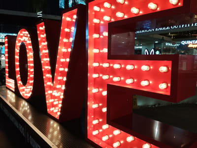 The image shows a large, illuminated "LOVE" sign in red with light bulbs. The sign is located in Playa del Carmen, Mexico, as indicated by the text "QUINTANA ROO" and "@QUINTANAROO" visible on the base of the sign. In the background, shops such as Starbucks and American Eagle Outfitters are visible through a glass partition. The scene appears to be at night, with the bright lights of the sign contrasting with the darker surroundings. There are no people clearly visible in the image. The sign is positioned on a metal textured platform.