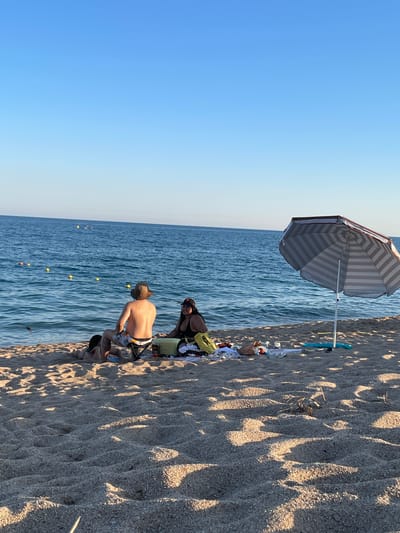 The image depicts a sunny day at the beach in Canet de Mar, Spain. The foreground is dominated by sand, with shadows from palm trees or umbrellas creating a dappled pattern. In the middle ground, two people are sitting on the sand near the water's edge. One person, wearing a hat and patterned swim trunks, is facing away from the camera, seemingly engaged in an activity on the sand. The other person, wearing a dark top, is seated next to them. A striped beach umbrella stands nearby, providing shade. Various beach items, including towels and bags, are scattered on the sand. The background features the calm blue sea, with a clear sky above. Yellow and orange buoys are visible in the water, marking a swimming area. The overall scene suggests a relaxed day at the beach, with the golden hour casting long shadows, indicating it is likely late afternoon. No visible text is present in the image.