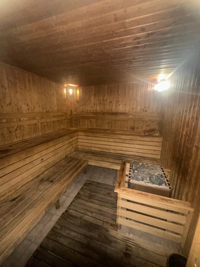 The image depicts the interior of a traditional wooden sauna. The walls, ceiling, and benches are all constructed from light-colored wood planks, creating a warm and natural aesthetic. Multiple tiers of benches line the perimeter of the room, offering ample seating. In the foreground, a sauna heater with stones is visible. A small, square window with a light fixture above it is located on the back wall. The floor appears to be a combination of wooden planks and tiles. The overall impression is that of a clean and well-maintained sauna facility. This scene is located in Samokov, Bulgaria.