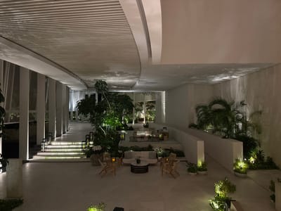 A spacious, modern outdoor lounge area features white tiled flooring and ample natural light. Lush greenery, including trees and palms, complements the minimalistic white architecture. Seating arrangements consist of sofas and chairs around coffee tables. Steps with lighting lead to a raised platform with additional seating. The ceiling has a partially curved, slatted design. The scene evokes a serene and luxurious ambiance.