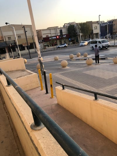 The image depicts a daytime street scene on a commercial thoroughfare in Riyadh, Saudi Arabia. The viewpoint is slightly elevated, looking across a paved pedestrian area towards a multi-lane asphalt road and buildings.
In the foreground, a dark metal handrail runs diagonally along the top of a light-colored concrete barrier. Below this, a concrete walkway descends. To the right of the walkway, three yellow and one black bollard are embedded in the ground.
The midground features a pedestrian crossing constructed from light and dark gray interlocking pavers. A series of seven light-colored spherical concrete bollards are arranged on this paved area, separating it from the vehicle traffic. Yellow and black striped crosswalk markings are visible on the street between some of these bollards. One male individual, wearing a dark shirt, dark trousers, and light-colored shoes, stands facing away from the camera on the paved pedestrian area, near a spherical bollard and the crosswalk, observing the street.
On the street, a white Toyota Hiace-style van is visible, moving from right to left. Behind it, a white sedan and a silver SUV are also present. The background consists of multiple commercial buildings lining the street. A prominent storefront displaying "ALRIFAI" (الرفاعي) with red accents is visible, likely a roastery or nuts shop. Other buildings are light-colored, multi-story structures. Dark-colored streetlights are interspersed along the street. The sky is clear and light blue, indicating daylight, with visible shadows suggesting the sun is relatively low.