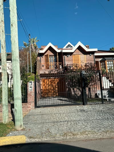 The image depicts a two-story brick house in Tigre, Argentina. The house features a unique roofline with multiple peaks and a triangular window on the top floor. Wooden shutters and frames adorn the windows. A black iron fence encloses the property. The street is paved with cobblestones, and a wooden fence runs along the sidewalk. Power lines are visible in the sky. The sky is clear and blue.