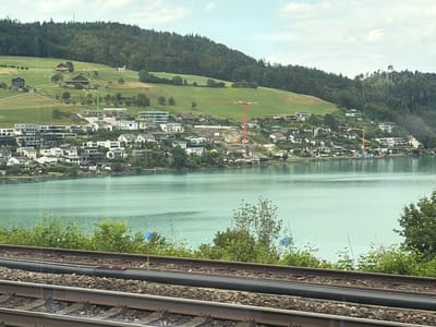 This media file captures a picturesque view of Immensee, Switzerland, featuring a lakeside town nestled against a verdant hillside. The foreground is dominated by a set of train tracks, indicating the perspective is from a moving train. Beyond the tracks, a tranquil, aquamarine lake stretches across the mid-ground, dotted with a few sailboats. The hillside behind the lake is covered in lush green grass and mature trees, with several houses and modern buildings dotting its slopes.
In the upper part of the hillside, traditional Swiss-style houses with dark roofs are visible, hinting at the area's history. Closer to the lake, contemporary architectural styles are evident in the multi-story buildings with clean lines and ample windows. A prominent feature on the hillside is a large red construction crane, suggesting ongoing development or renovation in the town. The sky above is partly cloudy, with patches of blue peeking through, suggesting a pleasant, possibly late spring or summer day. The overall atmosphere is serene and idyllic, showcasing a harmonious blend of nature and human habitation. No visible text is present in the image.