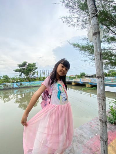 A young girl stands outside on what appears to be an outdoor park or recreation area in Kalitengah, Indonesia. She is wearing a pink dress. The background shows a waterway, likely a river or canal, with vegetation along the banks. In the distance, structures and power lines are visible, indicating the presence of a settlement or urban area.