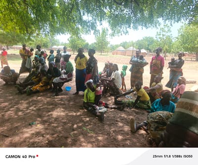 A gathering of women and children takes place on the ground in Gidan Kogi, Nigeria. The setting appears to be outdoors, with the group resting under the shade of trees. Buildings with thatched roofs are seen in the background. Many of the women are sitting or crouching, and some are holding infants or young children. Various garments in different colors and patterns are evident on the people. The ground is earthen with patches of vegetation and fallen leaves. A person wearing a hat is in the foreground.