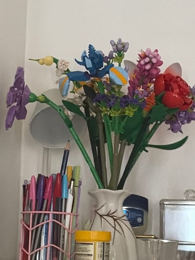 This media file shows a close-up view of artificial flowers in a vase, placed on a shelf. The flowers are a mix of colors, including red, pink, purple, blue, and yellow. Some of the flowers appear to be roses and hydrangeas. The vase is white with a delicate brown floral design. To the right of the vase, there is a stack of pens and pencils held together by a pink rubber band, and a yellow box, possibly containing more writing utensils or stationery. In the background, there is a reflective surface, possibly a mirror or a framed picture. The scene appears to be indoors, with neutral lighting suggesting daytime. The overall impression is one of a decorative display on a shelf, perhaps in a home or office setting. No people are visible. There is some text on the yellow box, but it is too blurry to read. The location context is Evere, Belgium.