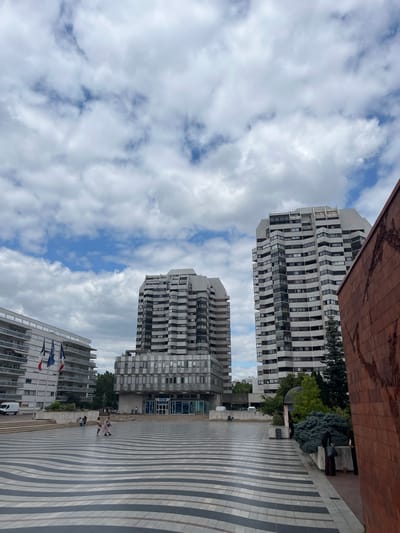 This media file shows an outdoor scene in Créteil, France, featuring modern architecture and a cloudy sky. In the foreground, a patterned pavement with alternating dark and light stripes leads towards two tall, white, multi-story buildings with numerous windows and balconies. The buildings have a distinctive stepped or curved facade.
In the middle ground, several people are visible. Some are sitting on a stepped area in front of the buildings, while others are standing and walking on the patterned pavement. There are also some trees and greenery interspersed with the buildings. A large, reddish-brown textured surface, possibly part of another building, is visible in the upper left corner, contrasting with the smooth facades of the residential towers.
In the background, more buildings and trees are visible, suggesting an urban environment. Several flags, including what appears to be the European Union flag and the French flag, are displayed on poles in front of one of the buildings. A white van is parked near the flags.
The sky is filled with a mixture of white and grey clouds, with patches of blue sky peeking through, indicating a partly cloudy day. The lighting suggests it is daytime, likely late morning or afternoon. The overall mood of the scene appears relaxed and casual, with people enjoying the outdoor space. No discernible text is visible in the image.