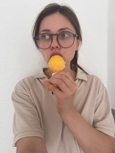 A woman with glasses is eating a yellow popsicle. The background is a plain white wall. The woman has light skin and brown hair. She is wearing a beige polo shirt. The location is unknown.