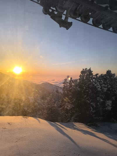 The image captures a snow-covered mountainous landscape at either sunrise or sunset within the Amirsay ski resort, Uzbekistan. A bright, low-angle sun on the far left horizon casts a strong golden light and visible lens flares across the scene. The sky transitions from a brilliant orange and yellow near the horizon to a clear, pale blue overhead. A distinct, uniform layer of white clouds or fog extends across the mid-ground, obscuring the valleys and giving the appearance of viewing from above the clouds.
Snow-covered coniferous trees, characterized by their dark green foliage partially covered in white snow, are prominent across the slopes in the mid-ground and right side of the frame. The immediate foreground consists of an undulating, pristine snow-covered slope, displaying subtle textural variations and long, dark shadows that extend from right to left. A slender, dark, vertical stick-like object protrudes from the snow in the lower right foreground. In the upper portion of the frame, specifically the top center and right, a dark, metallic mechanical structure is visible, comprising multiple wheels, axles, and a structural frame, consistent with the overhead track and cable assembly of a ski lift or gondola system. No individuals or specific activities are depicted.