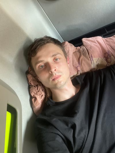 The media file shows a close-up, rotated, overhead shot of a young man lying down. He is wearing a black t-shirt and has light brown hair and green eyes. His expression is neutral, and he appears to be looking directly at the camera. He is lying on a patterned pink and brown pillow, which is placed on a light grey surface. To the right of the man, a portion of a grey object with a bright green interior is visible at the bottom right corner. The lighting suggests it is daytime, but no specific time of day can be determined. The setting is unclear, but the grey surface and the object on the right could be part of a vehicle or furniture. There is no visible text in the image. The man's relaxed posture and the soft lighting suggest a moment of rest or downtime.
