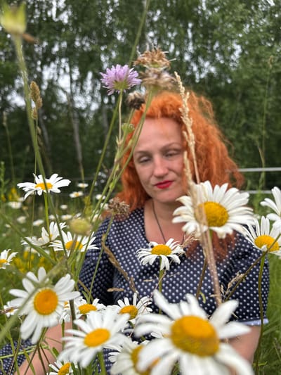 An out-of-focus, medium shot captures a woman with bright red hair and red lipstick in the foreground of a field of daisies. She is wearing a dark blue polka dot shirt and appears to be looking down, possibly at the flowers. The background is blurred, showing green trees and a hint of a white fence. The overall impression is one of a candid, outdoor portrait taken in a natural setting. The provided context indicates this image was taken in the city of Privolzhsky, Russia, suggesting a rural or semi-rural environment characteristic of the region.