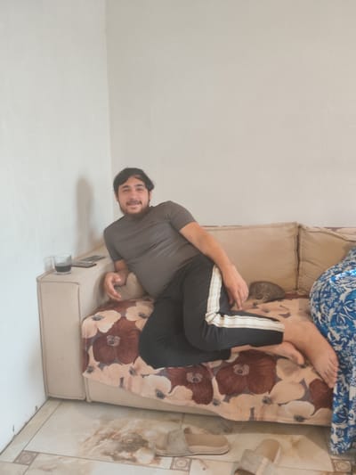 A man is lounging on a sofa in a room. He is wearing a grey t-shirt and black track pants with white stripes. He is smiling and looking towards the camera. Beside him on the sofa, a small kitten is sleeping. On the armrest of the sofa, there is a glass of dark liquid and two cell phones. The sofa is covered with a floral patterned blanket. The room appears to be a living room with light-colored walls and tiled flooring. A blue and white patterned fabric is visible on the right side of the sofa, possibly part of another piece of furniture or a throw. The scene suggests a casual and relaxed atmosphere. No specific time of day or weather is indicated. The location context is Temernitskiy, Russia.