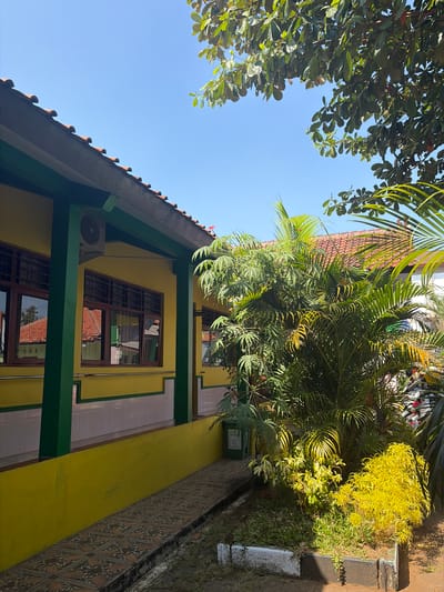 The image depicts the exterior of a building, likely in Pasekaran, Indonesia, under a clear blue sky. The building's facade is painted yellow with green trim. It features several windows with dark brown frames and latticed panes. An air conditioning unit is mounted on the exterior wall. A tiled pathway runs along the side of the building, bordered by a low yellow wall and a patch of grass. Lush green and yellow foliage, including palm trees, is prominent in the foreground and to the right of the building. The roof is covered with red tiles, and trees with large green leaves are visible at the top of the frame. The scene is illuminated by bright sunlight, casting shadows. There are no people visible in the image.
