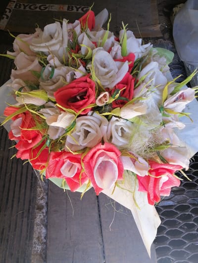 A bouquet of artificial flowers, predominantly red and white, is presented on a dark, weathered wooden surface. The bouquet is wrapped in light green and white paper, with some decorative green and yellow strands interspersed among the blooms. The background shows a dark surface with what appears to be lettering that reads "BYGG K BYGG," a grey plastic object on the right, and a dark metal grate or textured surface at the bottom right. The lighting suggests an outdoor or semi-outdoor setting, possibly a street or market stall in Lanivtsi, Ukraine, given the context provided. The overall impression is of a handmade craft item.