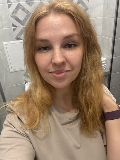 A young woman with blonde hair and freckles is taking a selfie in a bathroom. She is wearing a tan t-shirt and a purple watch on her left wrist. The bathroom walls are tiled with a geometric pattern. There is a white cabinet on the left side of the image and a toilet is visible in the background. The overall scene suggests a casual, personal moment.