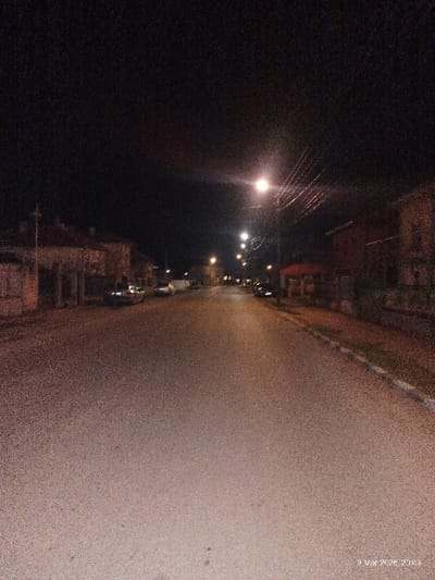 The image depicts a nighttime street scene in Voiniagovo, Bulgaria, captured on 9 March 2026, at 20:13. The asphalt road, appearing largely empty of active traffic or pedestrians, extends from the foreground into the distance under a dark sky. Multiple yellow-orange streetlights illuminate the scene from overhead poles, with visible utility wires. Residential-style buildings and houses line both sides of the street. On the left, several structures are visible behind fences, with at least two parked vehicles partially discernible near the curb. On the right, a sidewalk with a white and blue painted curb runs parallel to the street, bordering more buildings, one of which features a distinctive red-tiled roof. No individuals are present in the visible area.