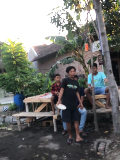 In the city of Putat, Indonesia, a group of young men is gathered outdoors. In the foreground, a young man in a black t-shirt and shorts holds a white plate. Behind him, another young man wearing a light blue t-shirt and blue shorts sits on a wooden bench. To the left, a third young man in a red and black plaid shirt is also seated. They are positioned in an area with dirt ground and some vegetation. A rustic wooden bench is a central object in the scene, with a pole on the right side of the frame. The background shows a house with a tiled roof and trees, suggesting a residential setting. The lighting indicates it may be late afternoon or early morning, with the sun casting a glow.