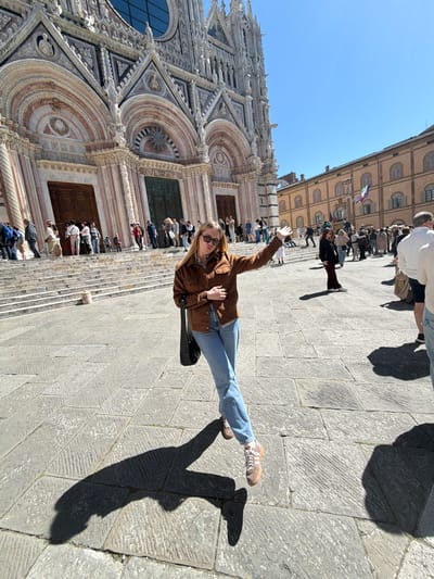 The image captures a vibrant, sunlit scene in an Italian city, specifically Siena, as hinted. On the left, the magnificent facade of a large, ornate cathedral, consistent with Siena Cathedral, stands tall. Its intricate Gothic architecture features detailed carvings, multiple arched entrances, circular windows, and decorative patterns in shades of white and reddish-brown stone.
In the foreground and extending to the right is a wide, sun-drenched plaza paved with large, irregularly shaped stone slabs. A young woman, the main subject, stands prominently in the mid-ground. She is smiling and waving towards the camera, wearing sunglasses, a brown jacket, light blue jeans, and light-colored sneakers. Her posture suggests a relaxed, happy mood. A black shoulder bag hangs by her side.
Numerous other individuals are visible in the background, milling about the plaza and ascending the steps leading to the cathedral's entrance. They appear to be tourists or sightseers, casually dressed. Across the plaza, a multi-story, light-colored building with numerous windows and closed shutters is visible in the background. An Italian flag, with vertical green, white, and red stripes, flies from this building.
The bright, clear blue sky indicates excellent weather. Distinct, elongated shadows cast by the people on the pavement suggest strong sunlight, likely during the morning or late afternoon. The overall scene depicts a lively day of sightseeing in a historic location. No discernible text is visible in the image.