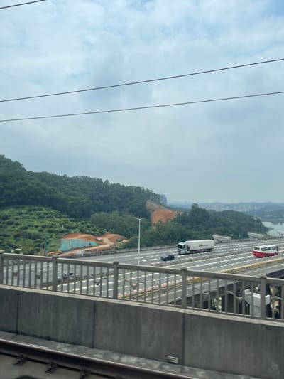 The image captures a view from a bridge overlooking a highway and a lush, green mountainside in Dongguan, China. The highway, with multiple lanes and white dashed lines, is busy with traffic, including a white truck with "TOWO" printed on its side and a red and white bus. The mountains are covered in dense vegetation, with an area of exposed earth and construction materials, possibly indicating ongoing development or land reclamation.
The sky is overcast, suggesting a cloudy or hazy day. The time of day is difficult to determine precisely, but the general lighting conditions imply it is daytime. The scene is one of a developed transportation corridor adjacent to a natural, mountainous landscape. The image is captured from a high vantage point, possibly from another elevated structure or a vehicle moving on the bridge.
Visible text includes "TOWO" on the side of a truck. The overall impression is a mix of urban infrastructure and natural scenery, characteristic of many rapidly developing regions. There are no people clearly visible as distinct subjects, though the presence of vehicles implies their activity. The mood is neutral, focusing on the visual elements of the scene.