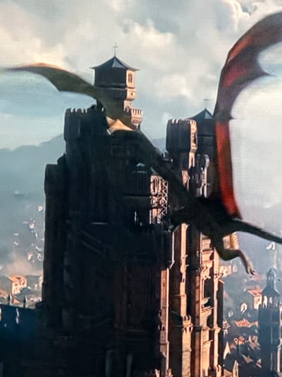 This is a digitally rendered image of dragons flying near a dark, imposing castle or fortress situated within a city. The castle has multiple towers with crenellated tops and distinct architectural features, including what appears to be a cross atop one of the spires. The dragons, one with a reddish underside to its wings, are in mid-flight, with one positioned in the foreground and another further back, partially obscured by the castle. The city below is depicted with numerous buildings, some with orange roofs, and a hazy, overcast sky above. The scene conveys a fantasy or epic setting. There is no discernible connection to the city of Izobilnyy, Russia, in this image.